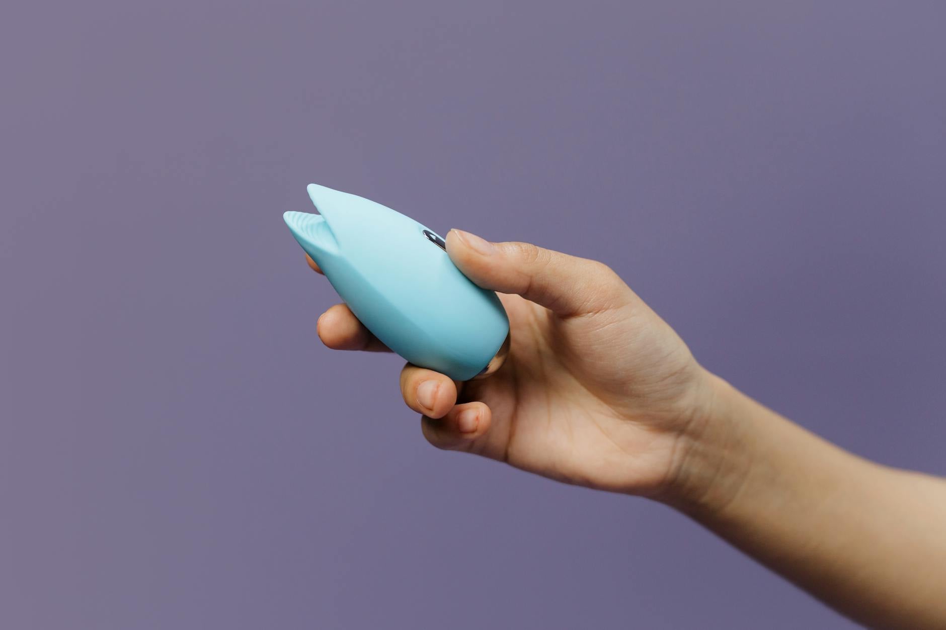 A hand holding a blue silicone lemon clitoral vibrator against a purple background